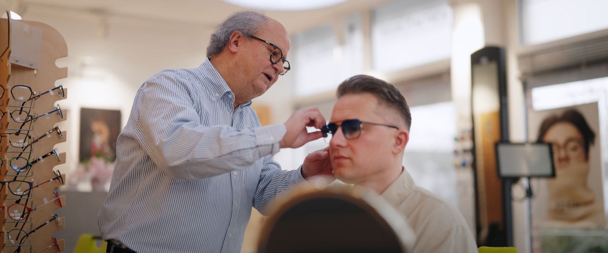 Herr Schünke hilft einem Kunden beim Anpassen einer neuen Brille. Sie stehen vor einem Spiegel im Verkaufsbereich, umgeben von Brillenständern.